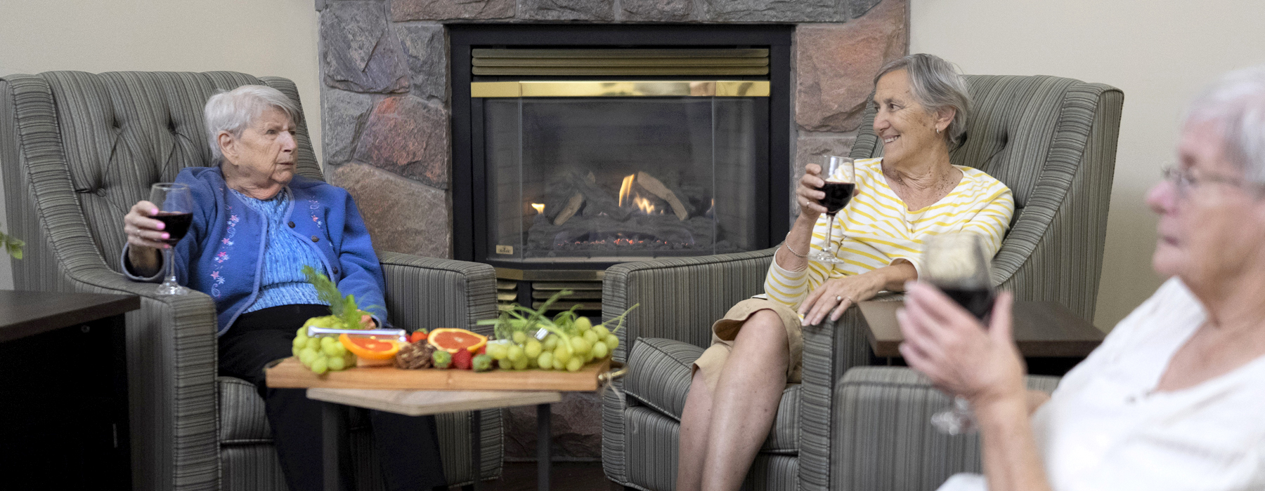 Resident Betty enjoying wine and cheese with friends by the fireplace in a cozy lounge at Atrium Retirement Residence in Orillia.